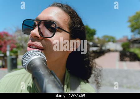 Wide angle close-up portrait of young Latino man with sunglasses, green clothes and long curly hair singing with microphone on street with sky in back Stock Photo