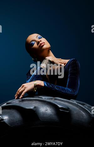 Young African American woman wearing a blue dress sits gracefully on top of a tire in a studio setting. Stock Photo