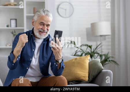 Joyful mature man with white hair jubilantly celebrates while looking at his smartphone, sitting comfortably in a living room setting. Stock Photo