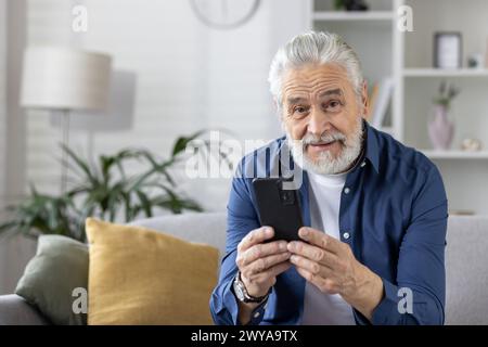 Cheerful elderly man with gray beard smiling as he looks at his smartphone, sitting comfortably on the couch at home. Stock Photo