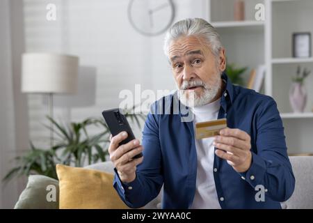 Elderly male with credit card and phone, potential victim of online fraud in a cozy home setting. Stock Photo