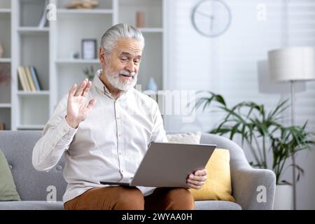 Cheerful elderly man having a video chat using a laptop from the comfort of his living room, waving hello. Stock Photo