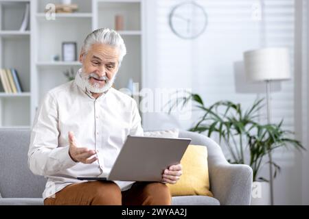 Elderly male using a computer in a bright, contemporary living room, staying connected and productive at home. Stock Photo