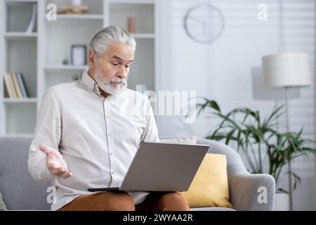 Focused senior male with a beard in a bright room, looking puzzled at a laptop while sitting on a couch. Stock Photo