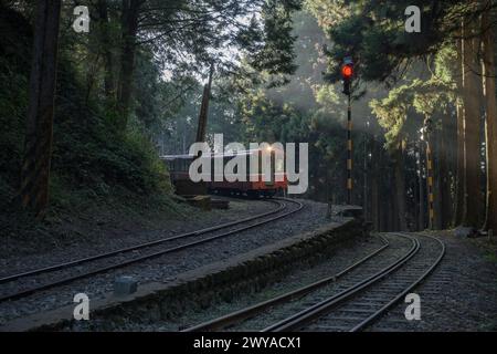 An old-fashioned train travels on a curved railway track through misty woods with light beams piercing through the forest in Alishan National Park Stock Photo