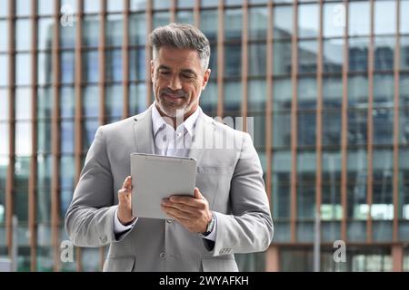 Happy middle aged businessman executive in suit using tablet standing outdoors. Stock Photo