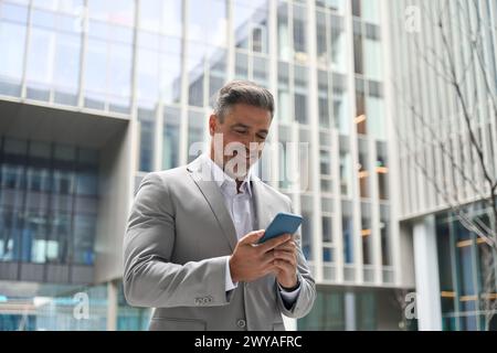 Happy mature businessman in suit using mobile phone standing outdoors. Stock Photo