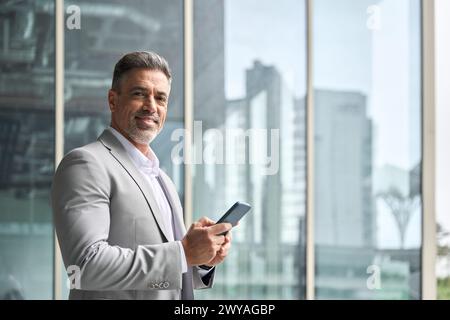 Middle aged businessman using phone standing at office window looking at camera. Stock Photo