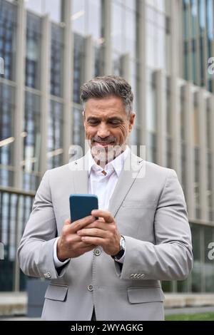 Happy middle aged business man ceo standing outdoors using smartphone. Stock Photo