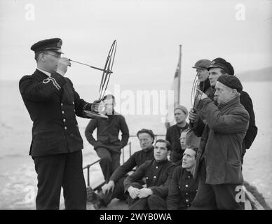 MOBILE DEMS INSTRUCTIONAL YACHT. 23 FEBRUARY 1942, ON BOARD THE MY MELITA, GOUROCK BAY. THE MOTOR LAUNCH IS THE FIRST INSTRUCTIONAL LAUNCH TO BE USED FOR THE DEFENSIVE EQUIPMENT OF MERCHANT SHIPS IN THE UNITED KINGDOM. SHE MAKES DAILY JOURNEYS TO THE ANCHORAGES TO KEEP THE MERCHANT SEAMEN INFORMED AND INSTRUCTED IN THE LATEST IMPROVEMENTS FOR THE DEFENCE OF THEIR SHIPS. - Merchant seamen undergoing instruction on board MY MELITA. 68 year old Moy Jones is seen holding the target finder. , Stock Photo