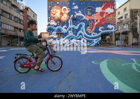 A young boy passes on a bicycle with his dog in front of a large, colorful dragon mural on a playground in Tainan city Stock Photo