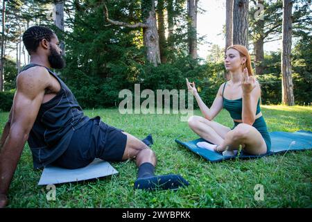 A man and a woman are sitting on the grass in a park. The man is wearing a black tank top and shorts, while the woman is wearing a green top and short Stock Photo