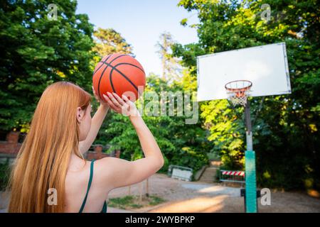 A woman is holding a basketball and shooting, sporty and healthy lifestyle. The scene is set in a park with trees and benches Stock Photo