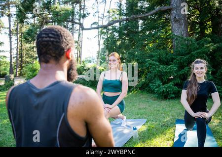 Three people are practicing yoga in a park. One of them is a man with a beard. The other two are women Stock Photo