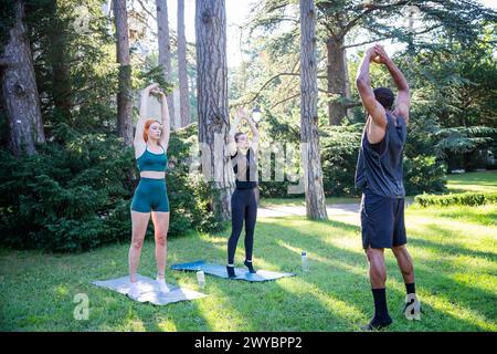 Three sporty people are exercising in a park doing some stretching exercises. Stock Photo