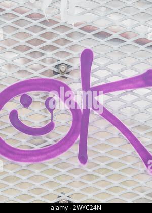 A close-up view of a section of metallic mesh with a vibrant purple graffiti drawing. The drawing consists of a stylized smiley face and abstract lett Stock Photo