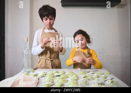 Two cute kids making dumplings in the home kitchen, dressed in beige chef's aprons. People. Culinary. Child learning cooking. Childhood. Domestic life Stock Photo