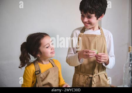 Authentic portrait of two adorable smiling kids making homemade dumplings at home kitchen, standing against white wall background Stock Photo