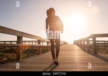 Rear view of a sporty woman carrying an exercise mat, walking along a boardwalk in the sun Stock Photo