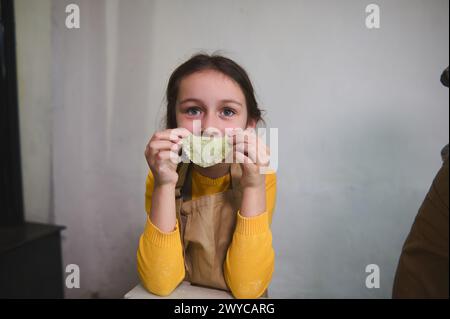 Portrait of a cute mischievous little girl in chef apron, holding a molded dumpling at her mouth level, making a beautiful smile looking at camera, st Stock Photo