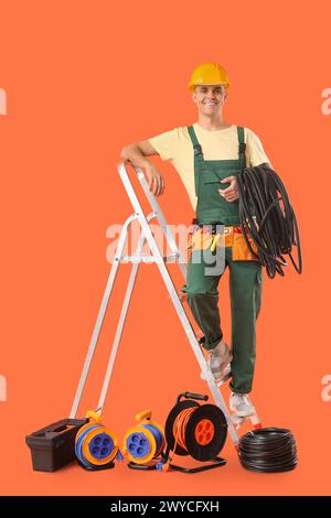 Young male electrician with flexible conduit tube and stepladder on orange background Stock Photo