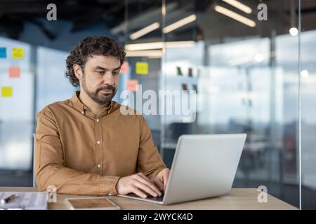 Focused professional man using a laptop in a contemporary office setting with glass partitions and sticky notes in the background. Stock Photo
