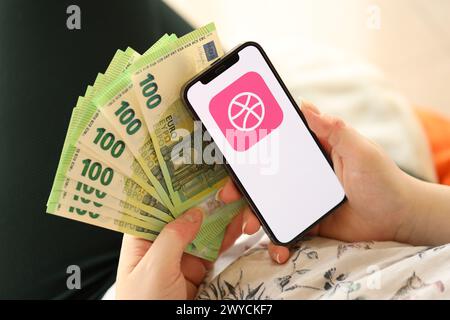 KYIV, UKRAINE - APRIL 1, 2024 Dribbble icon on smartphone screen and money in female hand. iPhone display with app logo and hundred euro bills in girls hands close up Stock Photo