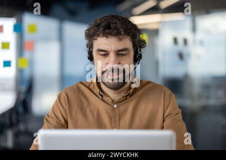 Focused Hispanic man using a headset and working diligently on his laptop at a modern office workspace. Stock Photo