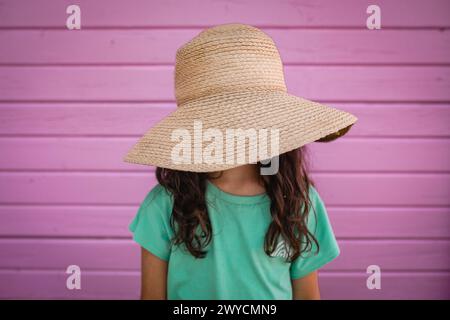 portrait of little girl hiding her face under big straw hat against bright pink wall Stock Photo