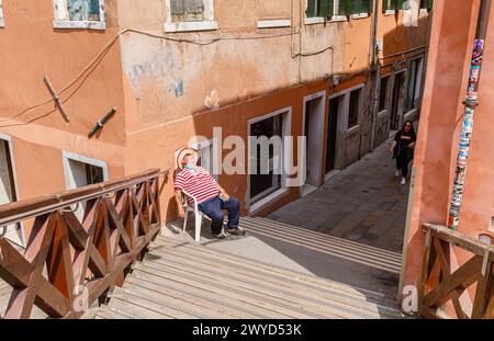 A gondolier in a traditional red and white hooped shirt sits resting in a chair asleep by Ponte Vinanti in Dorsoduro and Santa Croce, Venice, Italy Stock Photo