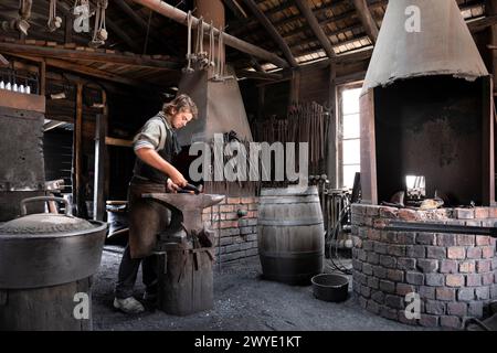 Blacksmith strikes ironwork (motion blur) on anvil with hammer in an old historic forge at the G.M Dilges' blacksmith shop Stock Photo