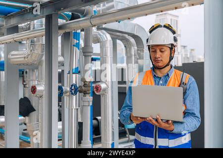 engineer man in a safety vest is standing around gas plump pipe, looking at a laptop. He is wearing a hard hat and a helmet Stock Photo