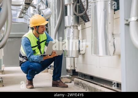 A man in a yellow vest is looking at a laptop while kneeling down. He is wearing a hard hat and safety glasses Stock Photo