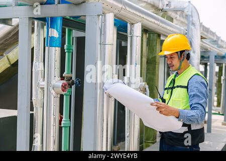 A man in a yellow helmet and a green vest is looking at a piece of paper. He is wearing a hard hat and he is a worker Stock Photo