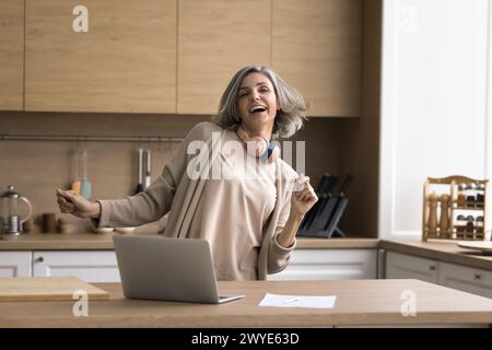 Happy mature woman enjoying music in kitchen at home Stock Photo