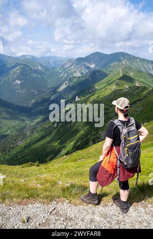 a woman looks down from the top of a mountain, dressed in hiking clothes, a red raincoat already tied around her waist, a backpack on her shoulders Stock Photo