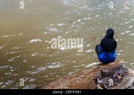A woman in a full-body swimsuit sits on the shore of a lake Stock Photo