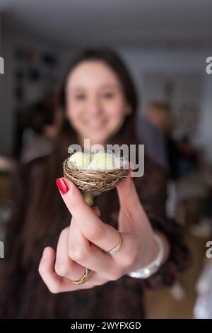 A smiling young woman holds up to the camera a small nest with three small chocolate Easter eggs painted green Stock Photo