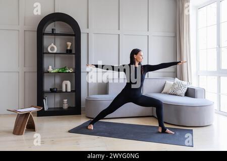 Athletic brunette practicing asanas at home, doing exercise Virabhadrasana 1, warrior pose number one, practicing yoga in black sports overalls standi Stock Photo
