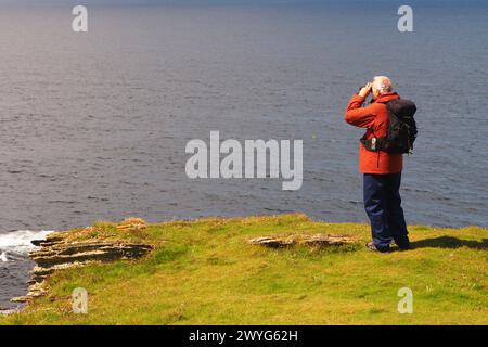 An older man 60+, wearing a red coat and black backpack standing on the cliffs on the Brough of Birsay, Orkney, Scotland UK, admiring the view Stock Photo