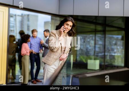 A stylish woman converses on her smartphone, walking past colleagues gathered in casual conversation Stock Photo