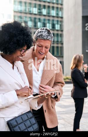 Multiracial mature business women couple gathered looking smiling at tablet, sharing work break. Stock Photo