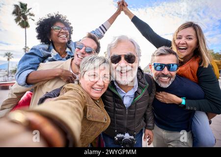 Group diverse funny mature tourist friends posing piggyback with hands joined in air taking selfie. Stock Photo