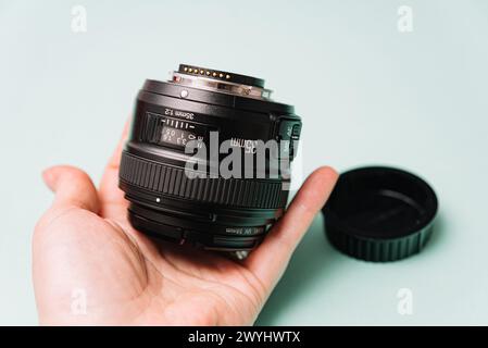 Close-up of a photographer's hand holding a black DSLR camera lens with a cap off against a soft blue background. Stock Photo