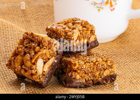Closeup view of crispy homemade peanuts, almond and chocolate mini protein bars and white coffee cup on sack cloth. Stock Photo