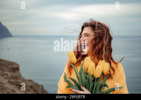 Portrait of a happy woman with hair flying in the wind against the backdrop of mountains and sea. Holding a bouquet of yellow tulips in her hands Stock Photo