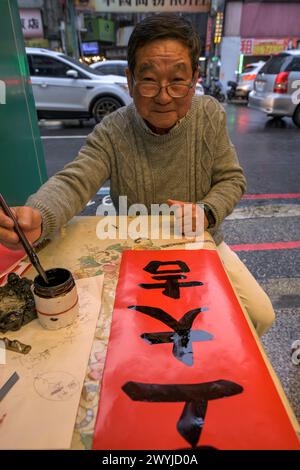 An elderly man focuses intently on writing Chinese characters with a brush on red paper, showcasing the art of calligraphy Stock Photo