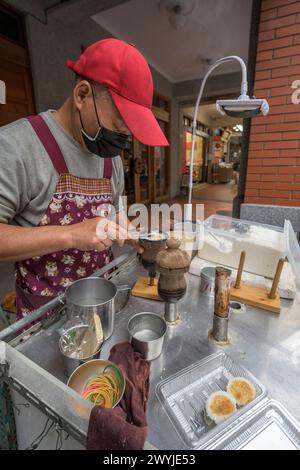 A street vendor wearing a mask attentively makes traditional desserts outdoors using a specialized utensil, during Chinese new year market Stock Photo