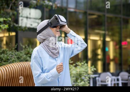 A joyful Muslim woman with a hijab using a virtual reality headset in an urban setting, showing excitement and curiosity. Stock Photo