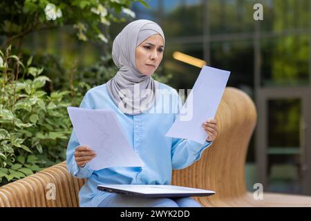 A focused professional woman in a hijab examines papers, sitting on an outdoor bench in a tranquil setting. Stock Photo